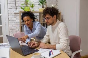 Man and woman sitting at a table pointing at a laptop screen.