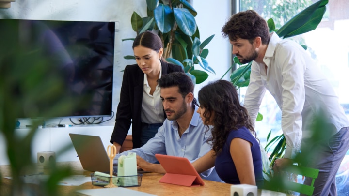 Man sitting at a table in an office looking at a laptop while two women and a man look at the laptop over his shoulder