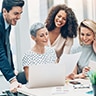 Woman sitting at a table holding a paper with a laptop in front of her while two women and a man lean over and read the paper.