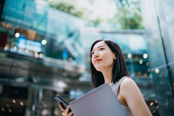 Asian woman with tablet and cellphone standing in front of modern glass building