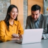 A businesswoman and businessman sitting at a table smiling while looking at a laptop