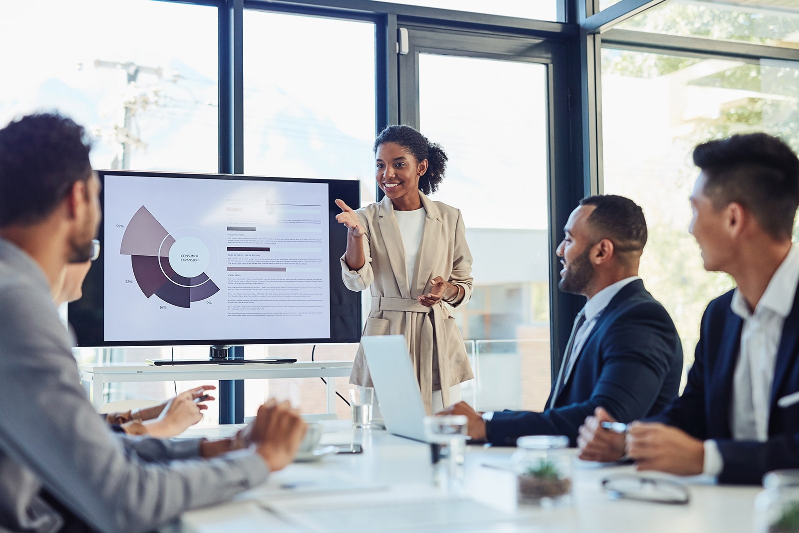 Young professional woman standing up presenting a slide to a table of male coworkers.